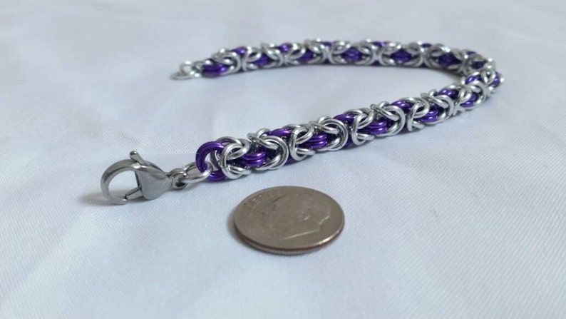 May include: A handmade bracelet featuring a Byzantine chain design. The bracelet is made of silver-colored metal links with purple accents. A silver clasp is attached. A dime is next to the bracelet.