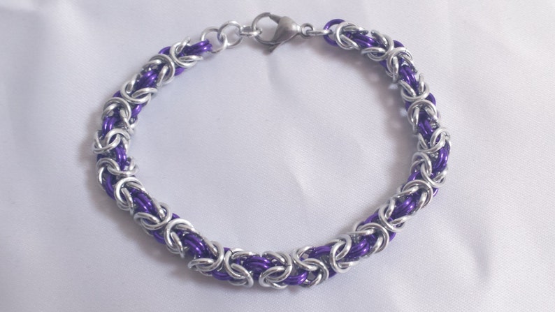 May include: A handmade bracelet featuring a chainmail design with alternating silver-tone and purple links. The bracelet has a lobster clasp closure and is displayed on a white surface. Jewelry.