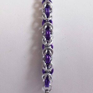 May include: A silver-colored chainmail bracelet with purple links. The bracelet features a series of interlocking metal rings, creating a textured and detailed design. The clasp is a simple, silver-toned hook.