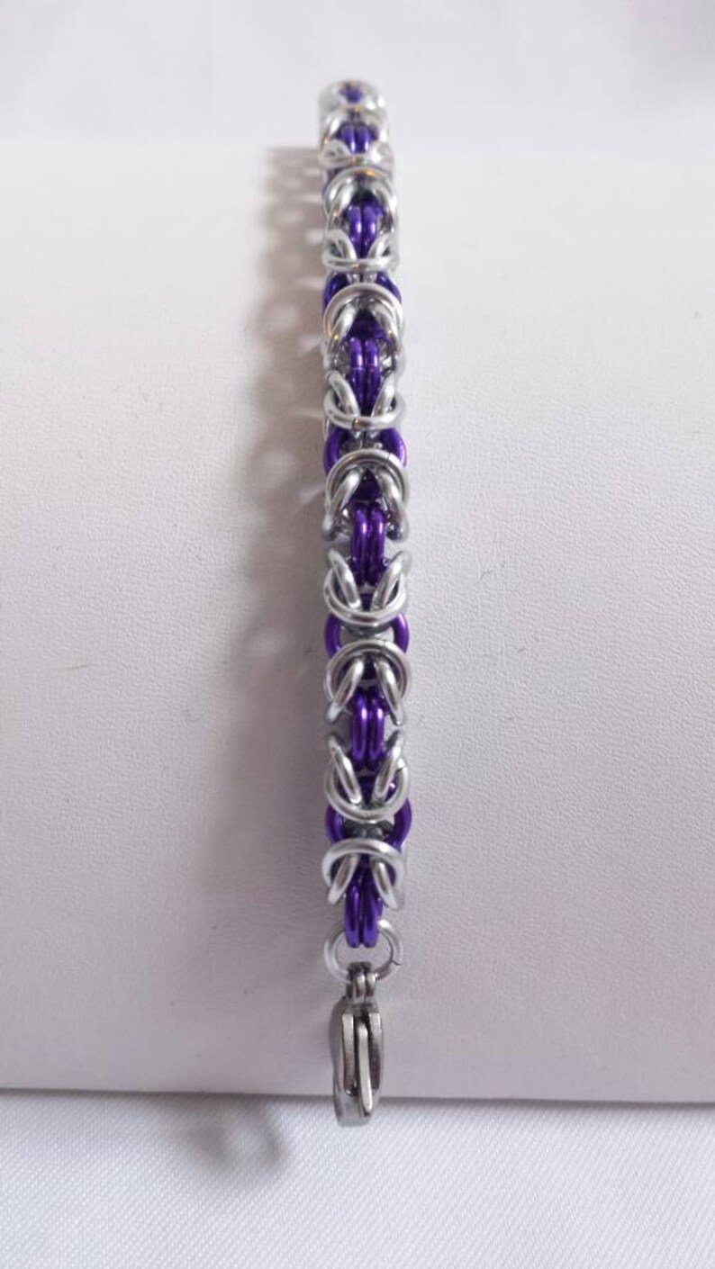 May include: A silver-colored chainmail bracelet with purple links. The bracelet features a series of interlocking metal rings, creating a textured and detailed design. The clasp is a simple, silver-toned hook.
