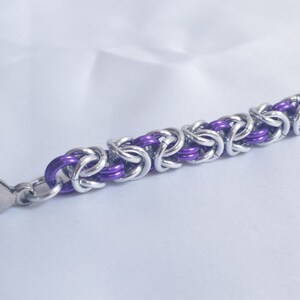 May include: A close-up of a silver and purple chainmail bracelet. The bracelet features a Byzantine weave pattern with alternating silver and purple links. A silver clasp is attached to the end of the bracelet.