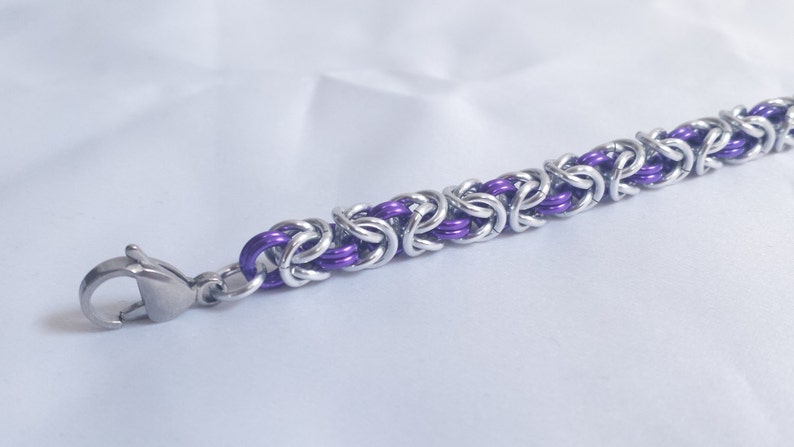 May include: A close-up of a silver and purple chainmail bracelet. The bracelet features a Byzantine weave pattern with alternating silver and purple links. A silver clasp is attached to the end of the bracelet.