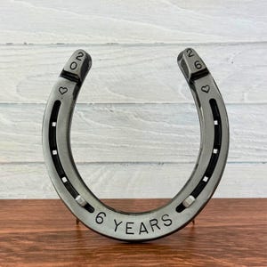 May include: A silver-toned metal horseshoe featuring stamped text and symbols. The left side displays the number 20 and a heart, while the right side shows 26 and a heart. The bottom curve is stamped with the text 6 YEARS.