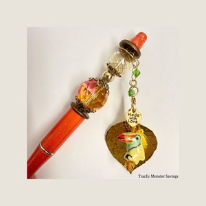 May include: Orange pen with gold and crystal embellishments. A gold leaf charm with a toucan and a heart charm with the text "Made with Love" hangs from the pen.