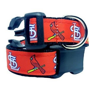 May include: A red pet collar featuring a repeating pattern of the St. Louis Cardinals logo and a cardinal bird perched on a yellow baseball bat. The collar includes a black plastic side-release buckle and a silver-toned metal D-ring.