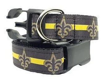 Könnte beinhalten: Ein schwarzes Haustierhalsband mit einem horizontalen gelben Streifen und einem wiederkehrenden goldenen Fleur-de-Lis-Muster. Das Halsband verfügt über eine schwarze Kunststoff-Steckschnalle und einen silberfarbenen D-Ring aus Metall.