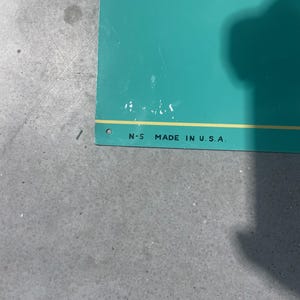 K&ouml;nnte beinhalten: T&uuml;rkisfarbenes Schild mit dem Text "N-5 MADE IN U.S.A." in Schwarz gedruckt. Das Schild hat einen gelben Streifen entlang der Unterkante und ein kleines Loch in der oberen linken Ecke. Das Schild liegt auf einer grauen Oberfl&auml;che.