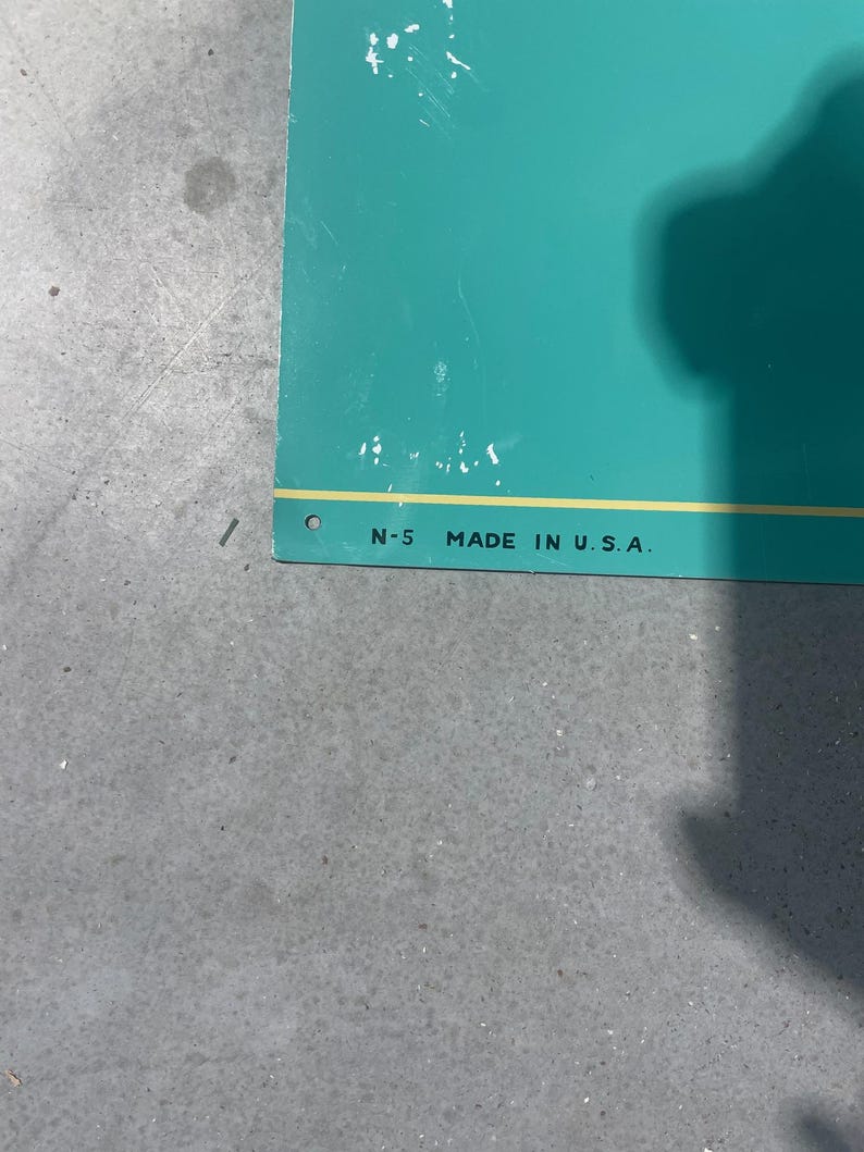 K&ouml;nnte beinhalten: T&uuml;rkisfarbenes Schild mit dem Text "N-5 MADE IN U.S.A." in Schwarz gedruckt. Das Schild hat einen gelben Streifen entlang der Unterkante und ein kleines Loch in der oberen linken Ecke. Das Schild liegt auf einer grauen Oberfl&auml;che.