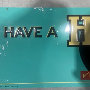 K&ouml;nnte beinhalten: Ein Vintage-Schild in T&uuml;rkis mit den Worten "HAVE A" in schwarzen Buchstaben und einem gro&szlig;en goldenen Buchstaben "H". Das Schild hat einen gelben Rand und den Text "MADE IN U.S.A." unten. Das Schild ist abgenutzt.