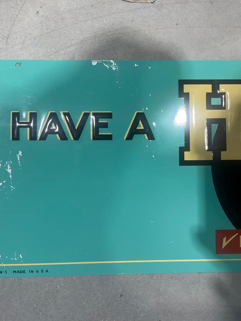 K&ouml;nnte beinhalten: Ein Vintage-Schild in T&uuml;rkis mit den Worten "HAVE A" in schwarzen Buchstaben und einem gro&szlig;en goldenen Buchstaben "H". Das Schild hat einen gelben Rand und den Text "MADE IN U.S.A." unten. Das Schild ist abgenutzt.