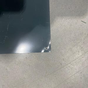 K&ouml;nnte beinhalten: Nahaufnahme eines dunkelgrauen, rechteckigen Objekts mit leicht reflektierender Oberfl&auml;che. Das Objekt weist leichte Kratzer und einen kleinen Chip an einer Ecke auf. Der Hintergrund ist eine hellgraue, strukturierte Oberfl&auml;che.