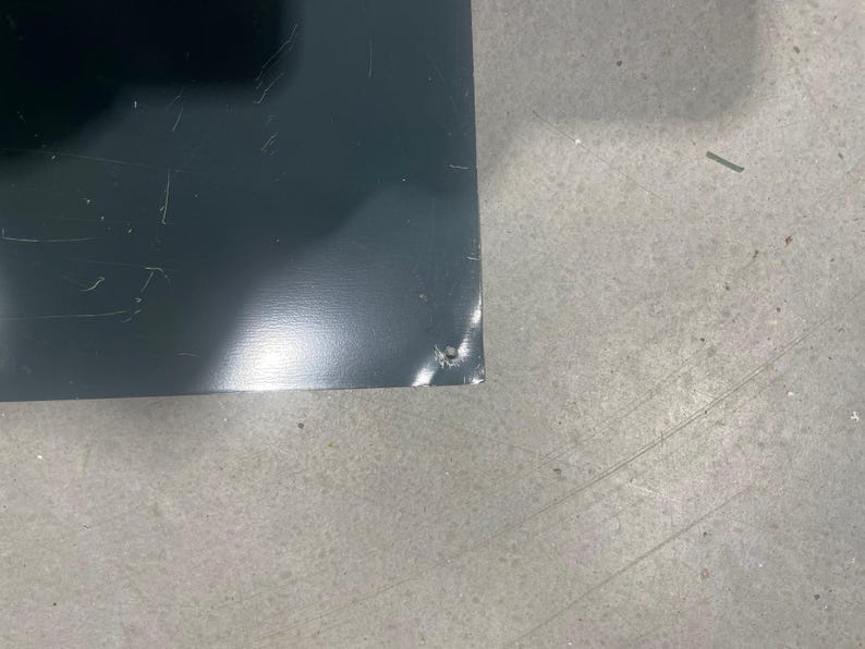 K&ouml;nnte beinhalten: Nahaufnahme eines dunkelgrauen, rechteckigen Objekts mit leicht reflektierender Oberfl&auml;che. Das Objekt weist leichte Kratzer und einen kleinen Chip an einer Ecke auf. Der Hintergrund ist eine hellgraue, strukturierte Oberfl&auml;che.