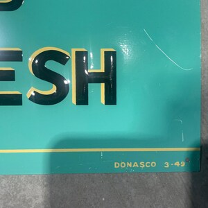 K&ouml;nnte beinhalten: Ein t&uuml;rkisfarbenes Schild mit dem Wort &bdquo;FRESH&ldquo; in gro&szlig;en, schwarzen Blockbuchstaben mit gelben Umrissen. Der Text &bdquo;DONASCO 3-49&ldquo; ist in kleinen, wei&szlig;en Buchstaben unten rechts aufgedruckt. Das Schild hat einen gelben horizontalen Streifen.