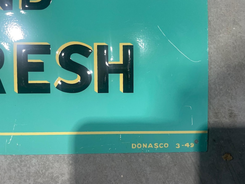 K&ouml;nnte beinhalten: Ein t&uuml;rkisfarbenes Schild mit dem Wort &bdquo;FRESH&ldquo; in gro&szlig;en, schwarzen Blockbuchstaben mit gelben Umrissen. Der Text &bdquo;DONASCO 3-49&ldquo; ist in kleinen, wei&szlig;en Buchstaben unten rechts aufgedruckt. Das Schild hat einen gelben horizontalen Streifen.