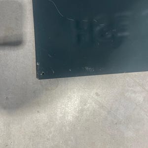 K&ouml;nnte beinhalten: Ein dunkelblaues rechteckiges Schild mit dem Teiltext "H&S" sichtbar. Das Schild weist einige Kratzer und Unvollkommenheiten auf. Der Hintergrund ist eine hellgraue, m&ouml;glicherweise betonierte Oberfl&auml;che.