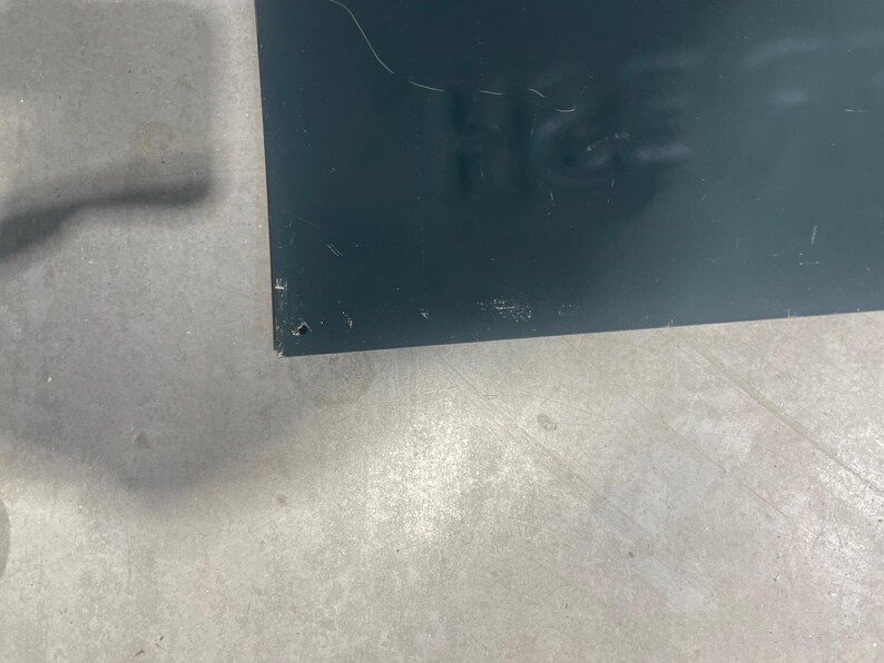K&ouml;nnte beinhalten: Ein dunkelblaues rechteckiges Schild mit dem Teiltext "H&S" sichtbar. Das Schild weist einige Kratzer und Unvollkommenheiten auf. Der Hintergrund ist eine hellgraue, m&ouml;glicherweise betonierte Oberfl&auml;che.