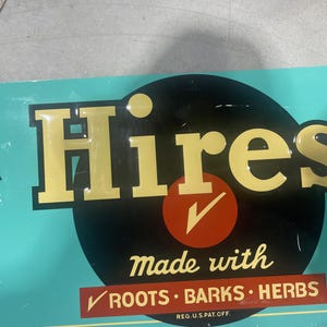 K&ouml;nnte beinhalten: Vintage Hires-Schild mit goldfarbenen Buchstaben auf einem schwarzen Kreis, vor t&uuml;rkisfarbenem Hintergrund. Das Schild lautet "Hires" mit "made with roots, barks, herbs" darunter. Ein roter Kreis mit einem H&auml;kchen ist ebenfalls vorhanden.