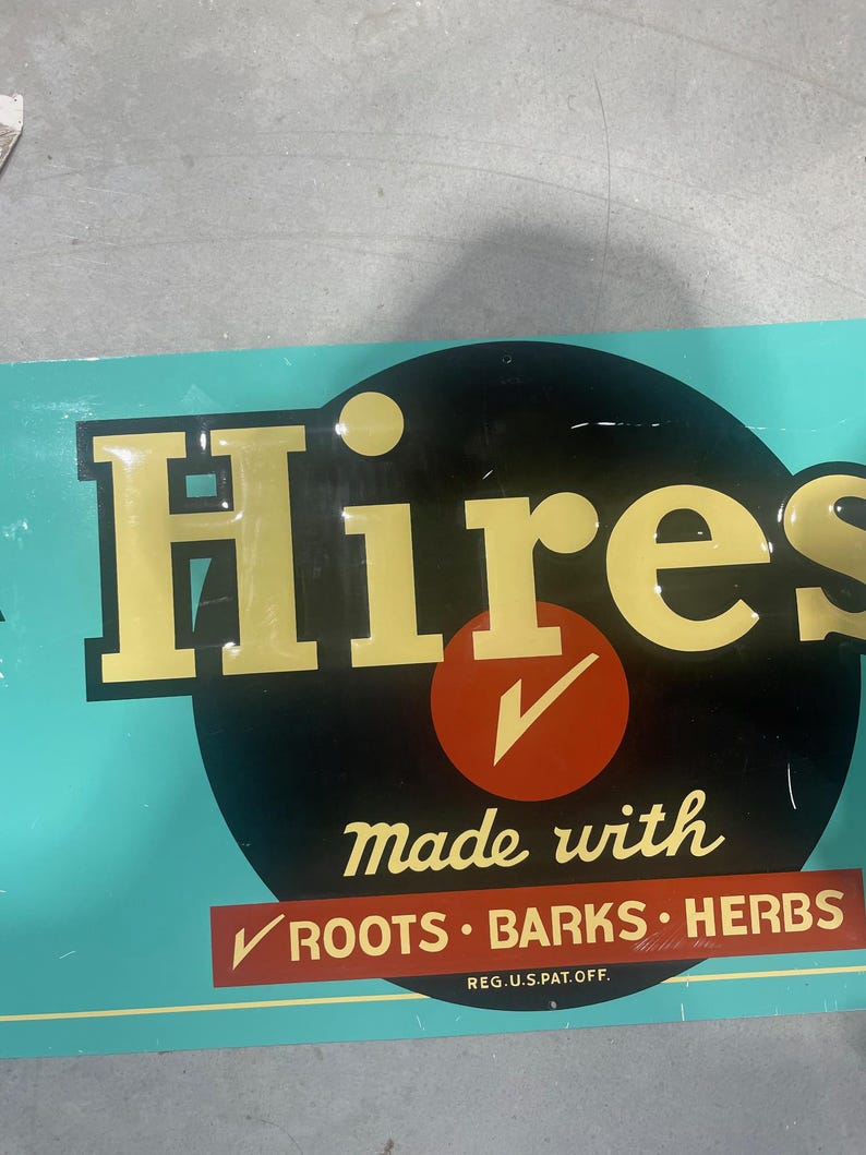 K&ouml;nnte beinhalten: Vintage Hires-Schild mit goldfarbenen Buchstaben auf einem schwarzen Kreis, vor t&uuml;rkisfarbenem Hintergrund. Das Schild lautet "Hires" mit "made with roots, barks, herbs" darunter. Ein roter Kreis mit einem H&auml;kchen ist ebenfalls vorhanden.