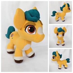 Puede incluir: Un peluche de pony en tonos amarillo, blanco, verde azulado y marrón. El pony tiene grandes ojos marrones, una melena y cola verde azulado, y un diseño de herradura en el flanco. El juguete es suave y tierno, con un diseño de dibujos animados.