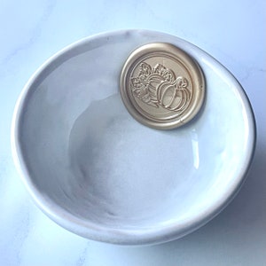 Puede incluir: Un tazón de cerámica blanco con un sello de cera dorado con un diseño de calabaza.