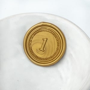 May include: A gold-colored wax seal with the number "1" in the center. The seal has a circular design with a beaded border and is set against a white background.