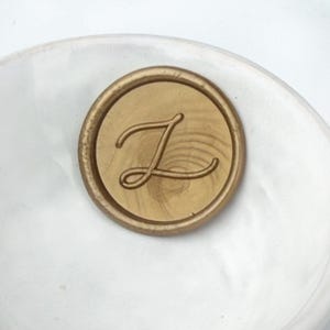 Pode incluir: Um selo de cera dourado com a letra "J" em uma escrita elegante. O selo é circular e tem uma textura de grão de madeira. O fundo é uma superfície branca desfocada.