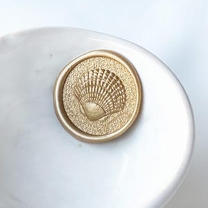 Puede incluir: Un sello de cera dorado con un diseño de concha. El sello es redondo y tiene un fondo texturizado. La concha está en el centro del sello. El sello está sobre una superficie blanca.
