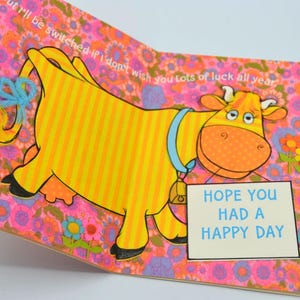 May include: A vintage greeting card featuring a cartoon cow with a yellow and orange body, blue accents, and a bell. The card has a floral background and text that reads "Hope You Had a Happy Day."