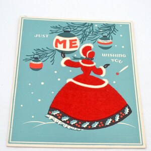 May include: A vintage Christmas card featuring a woman in a red dress with a white fur trim, holding a Christmas ornament that says "ME". The card has a blue background with white snowflakes and a green pine branch with ornaments.