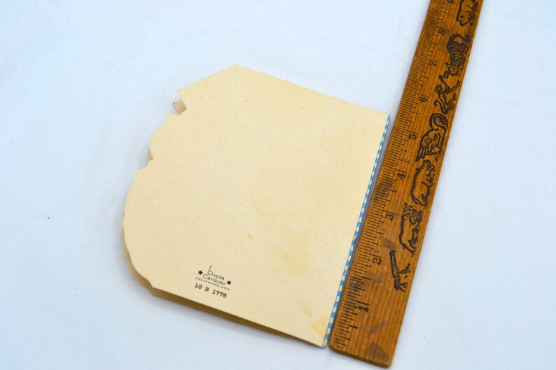 K&ouml;nnte beinhalten: Die R&uuml;ckseite einer Vintage-Gru&szlig;karte mit einem blau-wei&szlig; karierten Rand. Die Karte ist cremefarben und hat den Text "Buzza Card Co. 10 B 1776" in Schwarz gedruckt.