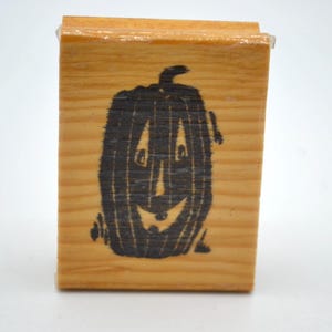 May include: A wooden stamp featuring a black jack-o'-lantern design. The pumpkin has a smiling face and is set against a light brown wooden block. The stamp is likely used for crafting or seasonal decorations.