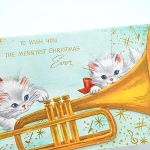 May include: A vintage Christmas card featuring two white kittens playing a golden trumpet. The card has a blue background with gold stars and musical notes. The text on the card reads "To wish you the merriest Christmas Ever."