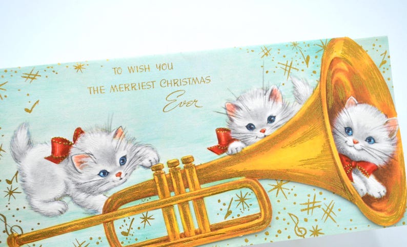 May include: A vintage Christmas card featuring two white kittens playing a golden trumpet. The card has a blue background with gold stars and musical notes. The text on the card reads "To wish you the merriest Christmas Ever."