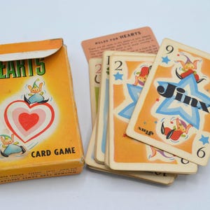 May include: Vintage Hearts 5 card game set. The box is yellow with a red heart and elf graphic. Several cards are visible, featuring the word "Jinx" and the numbers 2, 6, and 9. The game is designed with a whimsical, retro aesthetic.