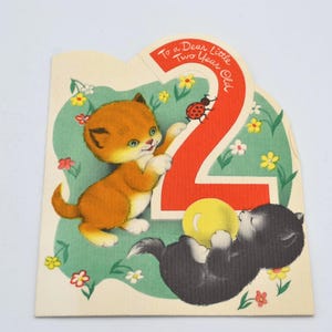 Puede incluir: Tarjeta de cumplea&ntilde;os vintage con dos gatitos de dibujos animados. Un gatito naranja est&aacute; de pie y el otro gatito negro est&aacute; acostado. La tarjeta tiene un n&uacute;mero 2 rojo y el texto "To a Dear Little Two Year Old."