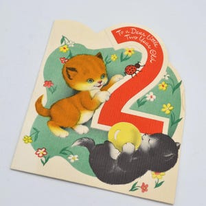 Puede incluir: Tarjeta de cumplea&ntilde;os vintage con dos gatitos y el n&uacute;mero 2. Un gatito naranja se acerca al n&uacute;mero, mientras que un gatito negro juega con una pelota amarilla. La tarjeta tiene un fondo verde con flores y el texto "To a Dear Little Two Year Old."