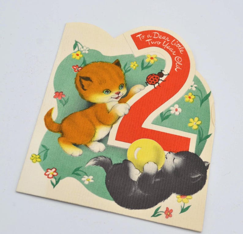Puede incluir: Tarjeta de cumplea&ntilde;os vintage con dos gatitos y el n&uacute;mero 2. Un gatito naranja se acerca al n&uacute;mero, mientras que un gatito negro juega con una pelota amarilla. La tarjeta tiene un fondo verde con flores y el texto "To a Dear Little Two Year Old."