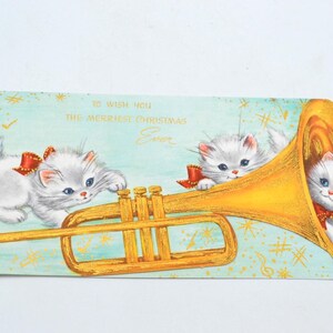 May include: A vintage Christmas card featuring two white kittens playing a golden trumpet. The card has a blue background with gold stars and musical notes. The text on the card reads "To wish you the merriest Christmas Ever."