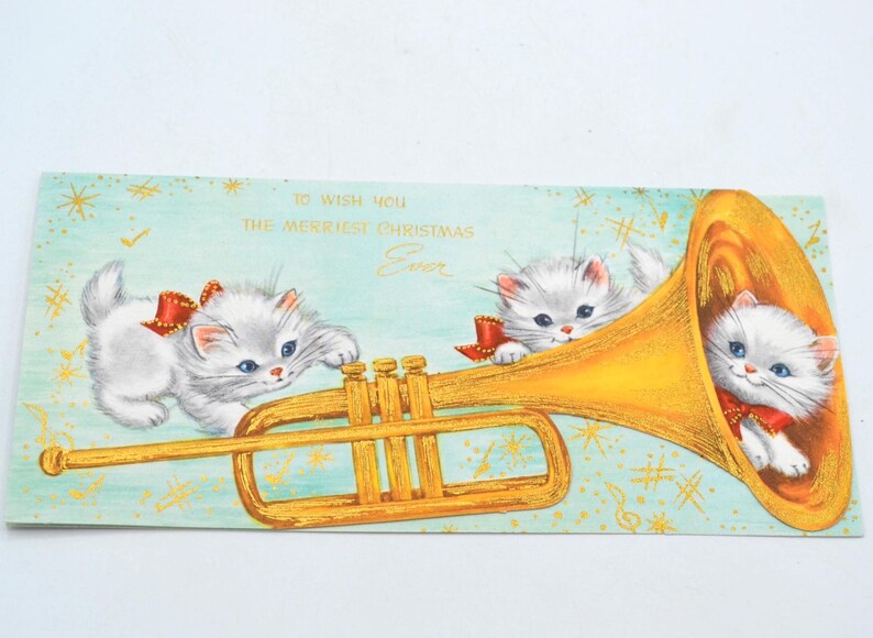 May include: A vintage Christmas card featuring two white kittens playing a golden trumpet. The card has a blue background with gold stars and musical notes. The text on the card reads "To wish you the merriest Christmas Ever."