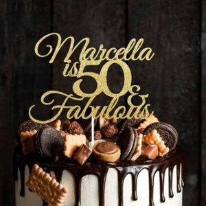 May include: A decorated birthday cake with a gold glitter cake topper that reads "Marcella is 50 & Fabulous." The cake has white frosting, chocolate drips, and is topped with cookies, chocolates, and candies. The cake sits on a wooden surface.