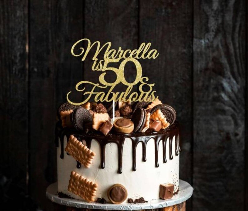 May include: A white frosted cake with chocolate ganache dripping down the sides. The cake is decorated with cookies, chocolates, and a gold glitter cake topper that reads "Marcella is 50 & Fabulous."