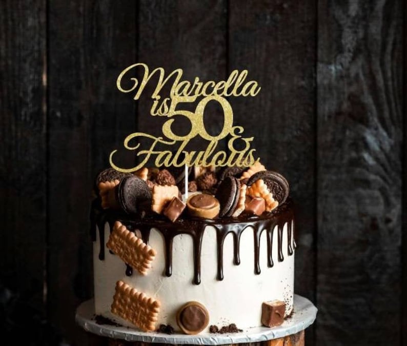 May include: A decorated birthday cake with a gold glitter cake topper that reads "Marcella is 50 & Fabulous." The cake has white frosting, chocolate drips, and is topped with cookies, chocolates, and candies. The cake sits on a wooden surface.