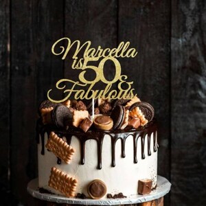 May include: A white frosted cake with chocolate ganache dripping down the sides. The cake is decorated with cookies, chocolates, and a gold glitter cake topper that reads "Marcella is 50 & Fabulous."