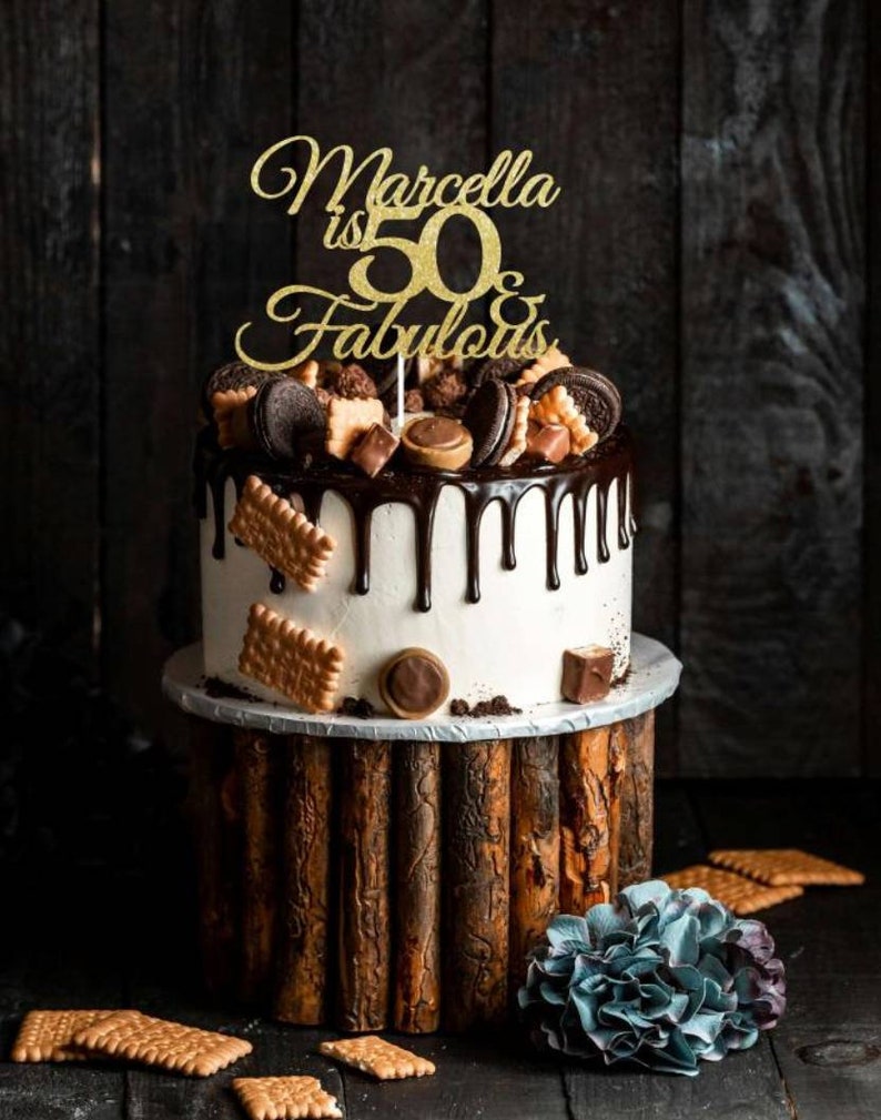 May include: A two-tiered cake with white frosting and chocolate ganache drips. The cake is decorated with cookies, chocolates, and a gold glitter cake topper that reads "Marcella is 50 & Fabulous". The cake sits on a wooden stand.