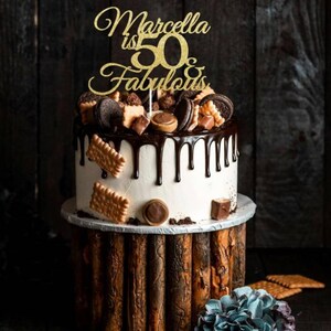 May include: A two-tiered cake with white frosting and chocolate ganache drips. The cake is decorated with cookies, chocolates, and a gold glitter cake topper that reads "Marcella is 50 & Fabulous". The cake sits on a wooden stand.