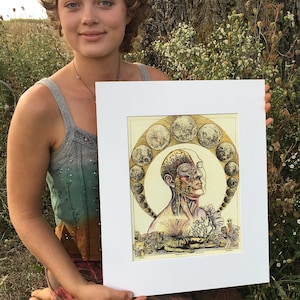 Op de afbeelding: Een vrouw houdt een ingelijst afdruk vast van een gedetailleerde anatomische illustratie van een menselijk hoofd met een cirkel van manen eromheen. De afdruk is op een crèmekleurige achtergrond met een gouden rand.