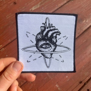 May include: A black and white embroidered patch featuring a stylized anatomical heart with an eye in the center. The heart is surrounded by a circle and radiating lines.