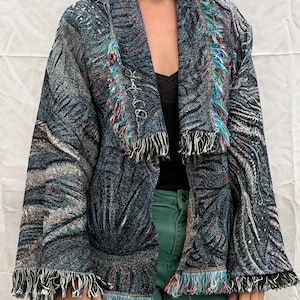 May include: A patterned jacket with a shawl collar and fringed edges. The jacket features a mix of blue, gray, and black colors with a textured design. The fringe includes red, blue, and white threads. The jacket is open in the front.