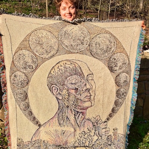 May include: Beige woven throw blanket featuring a detailed illustration of a human head with anatomical details, surrounded by a circular design of celestial bodies. The blanket has a fringed edge with blue, green, and red accents.