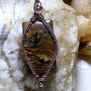 Peut inclure: Pendentif artisanal avec une pierre rectangulaire aux motifs bruns et jaunes, sertie dans du fil de cuivre. Le fil est finement tissé autour de la pierre, formant un cadre décoratif et une bélière. Le pendentif est présenté sur une surface texturée et claire. Le texte "Wild Raven Designs" est visible.